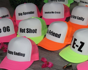 Puede incluir: Una colección de diez gorras de camionero blancas con paneles traseros de malla de color rosa neón, naranja y verde. Cada gorra tiene una frase diferente impresa en la parte delantera en letras negras. Las frases incluyen "The OG", "Hot Shot", "Happy Ending", "Big Spender", "Jamaica Me Crazy", "Ex-Friend", "Ice Ice Lolly", "E-Z", "Outnumbered", y "Fishin".