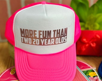 Puede incluir: Una gorra de camionero rosa y blanca con el texto "More Fun Than Two 20 Year Olds" en letras plateadas brillantes.