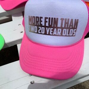 May include: Pink and white trucker hat with the text "More Fun Than Two 20 Year Olds" in gold lettering.