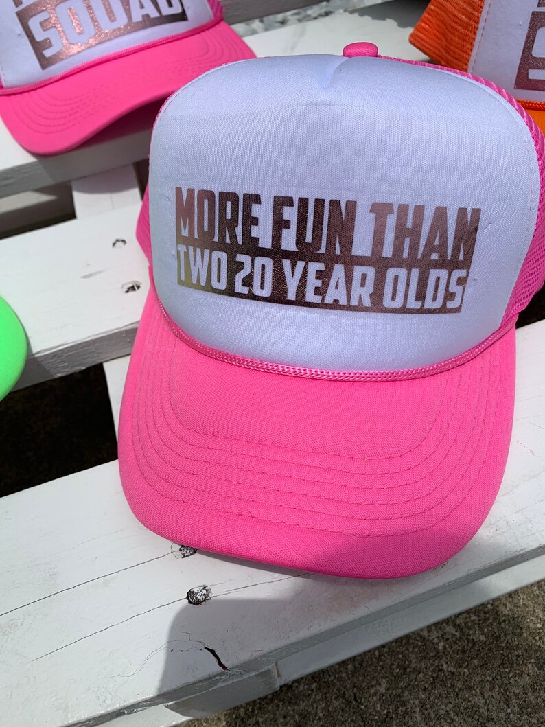 May include: Pink and white trucker hat with the text "More Fun Than Two 20 Year Olds" in gold lettering.