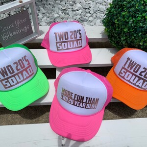 May include: Four trucker hats with a white front panel and a mesh back. The hats are in different colors: green, pink, orange, and hot pink. The front panel of each hat has a gold-colored graphic that says "TWO 20'S SQUAD". The pink hat has a different graphic that says "MORE FUN THAN TWO 20 YEAR OLDS".