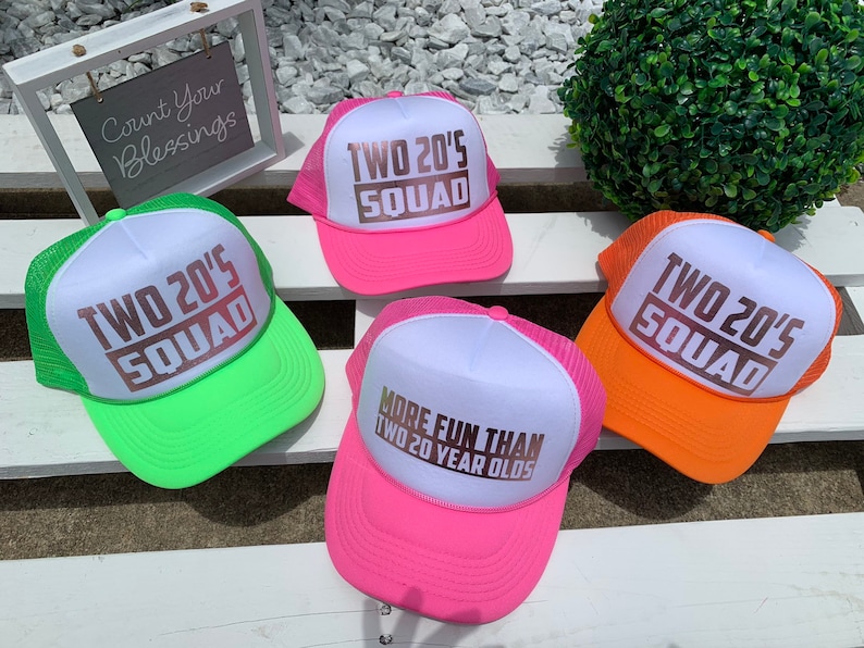 May include: Four trucker hats with a white front panel and a mesh back. The hats are in different colors: green, pink, orange, and hot pink. The front panel of each hat has a gold-colored graphic that says "TWO 20'S SQUAD". The pink hat has a different graphic that says "MORE FUN THAN TWO 20 YEAR OLDS".
