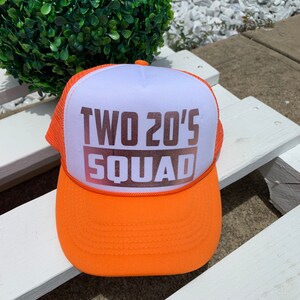 May include: Orange and white trucker hat with the text "TWO 20'S SQUAD" in brown letters.