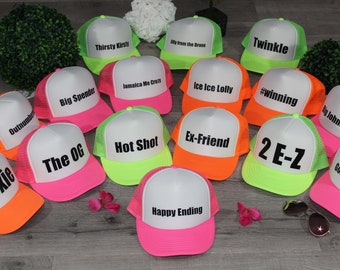 Puede incluir: Una colección de 15 gorras de camionero de neón con varias frases impresas en ellas. Las gorras son blancas con una malla de color rosa neón, naranja o verde en la parte posterior. Las frases incluyen "Thirsty Kirsti", "My from the Bronx", "Big Spender", "Jamaica Me Cray", "Ice Ice Lolly", "Outnumbered", "Foxie", "The OG", "Hot Shot", "Ex-Friend", "Happy Ending", "Twinkle", "#winning", "Big Johnson", "2 E-Z" y "Gone Fishin".