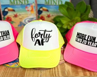 Puede incluir: Tres gorras de camionero con viseras de color rosa neón y amarillo. Las gorras tienen la parte de atrás de malla blanca y paneles frontales blancos con texto negro. Una gorra dice "forty AF", otra dice "More Fun Than Two 20 Year Olds", y la tercera gorra dice la misma frase.