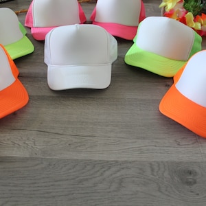 May include: Six blank white trucker hats with neon green, pink, and orange brims. The hats are arranged in a circle on a woodgrain surface.