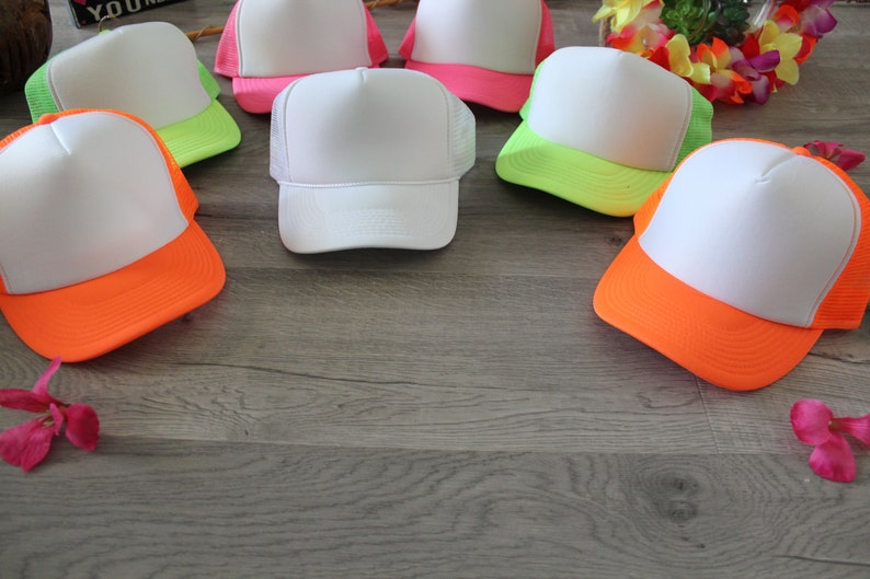 May include: Six blank white trucker hats with neon green, pink, and orange brims. The hats are arranged in a circle on a woodgrain surface.