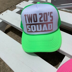 May include: A neon green and white trucker hat with the text "TWO 20'S SQUAD" in rose gold lettering.
