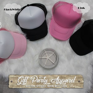 May include: Four trucker hats in different colors: black, white, pink, and pink and white. The hats are on a white fluffy surface. The text "Gift Party Apparel" is on a wooden sign below the hats.