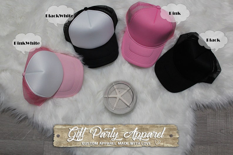 May include: Four trucker hats in different colors: black, white, pink, and pink and white. The hats are on a white fluffy surface. The text "Gift Party Apparel" is on a wooden sign below the hats.