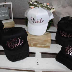 May include: Five black and white trucker hats with mesh panels. The hats have the words "Team Bride" written in pink script on the front. One white hat has the word "Bride" written in pink script on the front.