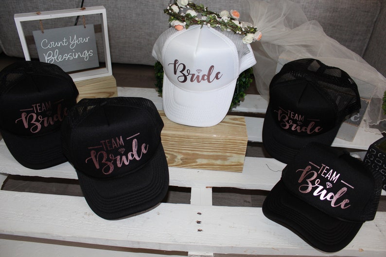 May include: Five black and white trucker hats with mesh panels. The hats have the words "Team Bride" written in pink script on the front. One white hat has the word "Bride" written in pink script on the front.