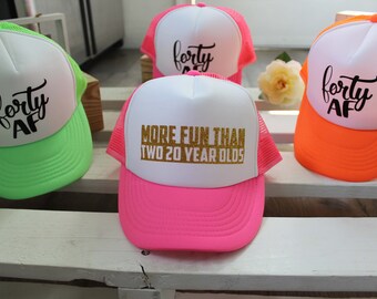 Puede incluir: Cuatro gorras de camionero con diferentes colores y diseños. Una gorra es rosa con malla blanca y dice "More Fun Than Two 20 Year Olds" en purpurina dorada. Otra gorra es verde neón con malla blanca y dice "Forty AF" en negro. Las otras dos gorras son blancas con malla negra y dicen "Forty AF" en negro.