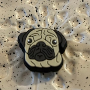 Pode incluir: Um pingente de sapato em forma de cabeça de cão pug com rosto bege claro, orelhas pretas e focinho preto. O pug apresenta rugas faciais detalhadas, olhos brancos redondos com pupilas pretas e uma área de nariz e boca plana e escura.