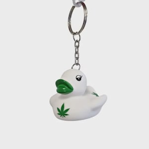 Puede incluir: Llavero de pato de goma blanco con labios verdes y un diseño de hoja de cannabis verde. El pato tiene una cadena y un llavero plateados. El llavero cuelga sobre un fondo blanco.