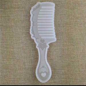May include: A clear comb-shaped mould with a handle featuring heart-shaped designs. The comb's teeth are visible, and the handle has a teardrop shape. The mould is made of a translucent material.
