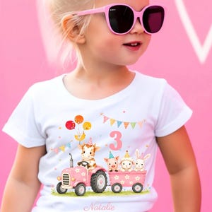 Könnte beinhalten: Weißes T-Shirt mit einer farbenfrohen Illustration eines rosa Traktors, der Tierfiguren transportiert, darunter eine Kuh, ein Schwein, ein Schaf und ein Kaninchen. Die Zahl "3" und der Name "Natalie" sind ebenfalls abgebildet. Das Kind trägt eine rosa Sonnenbrille.
