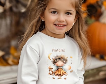 Könnte beinhalten: Ein junges Mädchen trägt ein weißes Langarm-Sweatshirt mit einer Feen-Grafik und dem Namen "Natalie" über der Grafik. Die Zahl "5" ist ebenfalls auf dem Sweatshirt aufgedruckt. Sie trägt eine Blumen-Haarschleife.