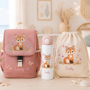 Könnte beinhalten: Ein altrosa Rucksack, eine weiße Trinkflasche und ein cremefarbener Kordelzugbeutel, jeweils mit einem Cartoon-Fuchs mit Blumenkranz, Schmetterlingen und dem in Schreibschrift gedruckten Namen Emily, die zusammen auf einer Fläche stehen.