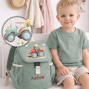Può includere: Uno zaino verde salvia con un disegno di trattore e il nome "Anton" ricamato in lettere colorate. Lo zaino è dotato di chiusura a patta, tasche laterali e fibbia. È presente anche un'illustrazione ad acquerello di un trattore.