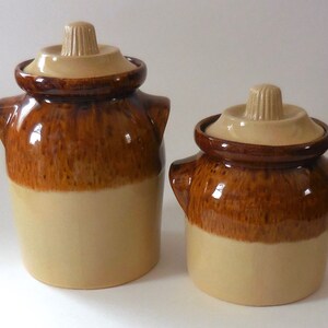 Könnte beinhalten: Zwei braune und beige Keramik-Gläser mit Deckeln. Das größere Glas befindet sich links und das kleinere rechts. Beide Gläser haben eine braune Glasur auf der oberen Hälfte und eine beige Glasur auf der unteren Hälfte.