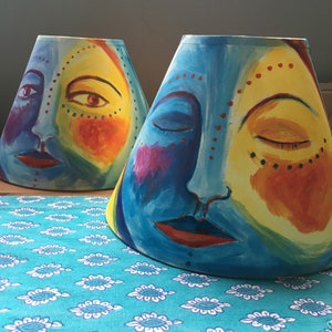 May include: Two lampshades with painted faces. The lampshades are blue, yellow, red, and purple. The faces have closed eyes and lips.