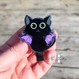 May include: A black cat with purple wings and green eyes. The cat is made of resin and has a glittery finish. It is a cute and whimsical charm.