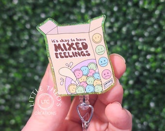 May include: A badge reel with a box design that reads "it's okay to have MIXED FEELINGS." The box contains colorful, round candies with various facial expressions. The reel has a clear plastic clip and a retractable cord.