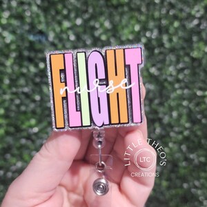 Op de afbeelding: Een kleurrijke badgehouder met de tekst "FLIGHT nurse" in regenboogkleuren op een zilveren glitterachtergrond. De badgehouder is bevestigd aan een intrekbare badgehaspel.