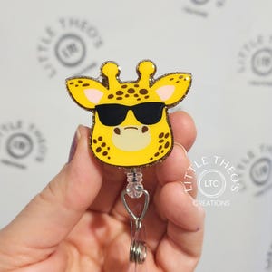 May include: A yellow glitter giraffe with black spots and sunglasses, attached to a retractable badge reel. The giraffe is wearing sunglasses and has a pink nose.