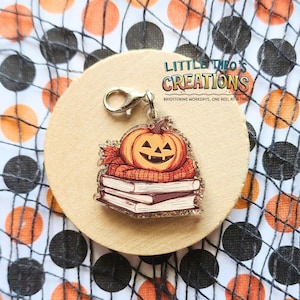 Op de afbeelding: Een Halloween-thema bedel met een lachende pompoen bovenop een stapel boeken, met een herfstkleurige sjaal. De bedel is bevestigd aan een zilverkleurige sluiting. De tekst "LITTLE THEO'S CREATIONS" is zichtbaar. De achtergrond heeft oranje en zwarte stippen.