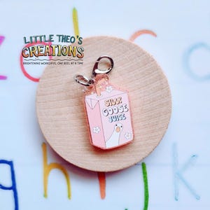 May include: A pink acrylic keychain shaped like a juice box, featuring the text "Silly Goose Juice" and a cartoon goose. The keychain has a silver clasp and is decorated with small flower details. The background is a wooden circle.