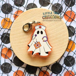 Op de afbeelding: Een Halloween-thema bedel met een spook verkleed als verpleegster, compleet met een medische tas, stethoscoop en verpleegsterskap. De bedel is bevestigd aan een zilverkleurige sluiting. De bedel ligt op een houten cirkel met de tekst "Little Theo's Creations".