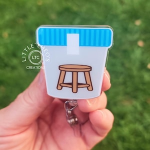 Puede incluir: Un porta credenciales de plástico a rayas blancas y azules con una imagen de dibujos animados de un taburete de madera marrón. El porta credenciales tiene un carrete retráctil transparente.