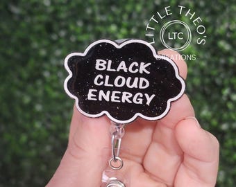 Può includere: Un porta badge a forma di nuvola nera con finitura glitterata e bordo bianco. Il testo BLACK CLOUD ENERGY è stampato in lettere maiuscole bianche sulla parte anteriore. Un cinturino in plastica trasparente e una clip in metallo sono fissati.