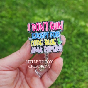 May include: A black glitter badge reel with the text "I don't run...except for code blue & ama papers" in colorful letters. The text "Little Theo's Creations" is printed below the badge reel.