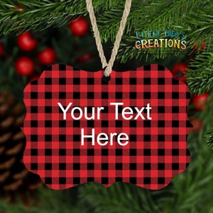 Puede incluir: Adorno navide&ntilde;o a cuadros rojos y negros con el texto "Your Text Here" en blanco. El adorno est&aacute; colgado de una cuerda de hilo natural. El fondo incluye agujas de pino y bayas rojas.