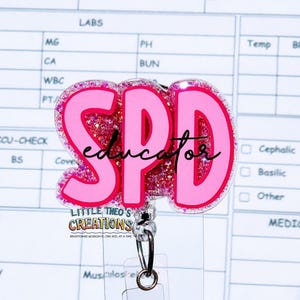 May include: A pink badge reel with the letters "SPD" in a bold font, with the word "educator" in cursive. The letters are outlined in glitter. The reel has a clear plastic holder and a metal clip.
