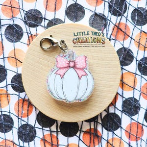 May include: A white pumpkin charm with a pink polka dot bow and silver clasp. The charm is on a wooden circle with the text "LITTLE THEO'S CREATIONS" and "BRIGHTENING WORKDAYS, ONE REEL AT A TIME". The background has a Halloween theme.