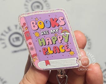 Puede incluir: Un portagafete en forma de libro rosa y morado con purpurina y el texto "BOOKS ARE MY HAPPY PLACE" en letras de colores. El portagafete está unido a un carrete retráctil de gafete plateado.