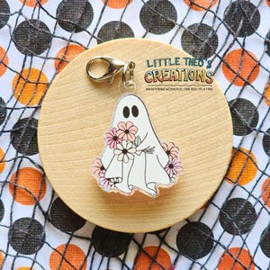 Op de afbeelding: Een witte spookvormige bedel met een boeket roze en witte bloemen, bevestigd aan een goudkleurige sluiting. De bedel ligt op een houten cirkel, met de tekst "LITTLE THEO'S CREATIONS" en "BRIGHTENING WORKDAYS, ONE REEL AT A TIME".