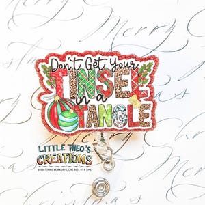 Puede incluir: Un portatarjetas festivo con la frase "Don't Get Your Tinsel in a Tangle". El diseño presenta un borde rojo brillante, adornos navideños y varios patrones dentro de las letras. El portatarjetas incluye un cordón retráctil y un clip metálico.