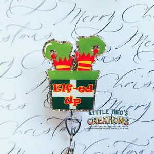 May include: A Christmas-themed badge reel with a green and gold glitter design. The badge features elf legs sticking out of a green box with the words "Elf-ed Up" in red and yellow. The reel has a silver clip and the text "Little Theo's Creations".