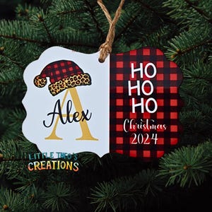 Puede incluir: Adorno navide&ntilde;o personalizado con borde festoneado blanco. El lado izquierdo presenta un dise&ntilde;o de gorro de Pap&aacute; Noel con borde de estampado de leopardo y el nombre "Alex" con una inicial dorada. El lado derecho tiene un patr&oacute;n de cuadros rojos y negros con "HO HO HO Christmas 2024".