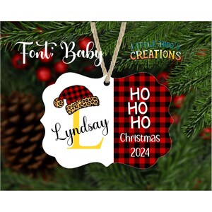 Puede incluir: Adorno navide&ntilde;o personalizado con borde festoneado blanco. El lado izquierdo presenta un gorro de Pap&aacute; Noel con borde de estampado de leopardo y el nombre "Lindsday" con una "L" amarilla. El lado derecho tiene un patr&oacute;n de cuadros rojos y negros con "HO HO HO Christmas 2024".
