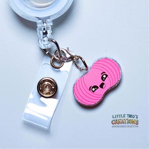 May include: A pink yarn ball badge reel with a cartoon face and silver glitter trim. The badge reel has a clear plastic strap and a metal clip. The text "LITTLE THEO'S CREATIONS" is visible.