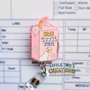 May include: A pink badge reel in the shape of a juice carton, featuring the text "SILLY GOOSE JUICE" and a cartoon goose. The reel has a glittery edge, a clear plastic snap, and a retractable cord. The text "LITTLE THEO'S CREATIONS" is also visible.