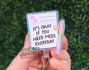 May include: A badge reel with a prescription pad design. The text reads "IT'S OKAY IF YOU NEED MEDS EVERYDAY." The badge has a glittery border and a pen graphic. The reel also features the text "LITTLE THEO'S CREATIONS".