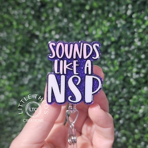 Könnte beinhalten: Ein lila-weißes Ausweis-Jojo mit dem Text "SOUNDS LIKE A NSP". Das Ausweis-Jojo hat ein glitzerndes Finish und einen transparenten Kunststoffclip. Der Text ist in einer fetten, serifenlosen Schriftart.
