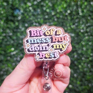 Könnte beinhalten: Ein glitzernder Badgehalter in Rosa, Blau, Gelb und Grün mit dem Text "Bit of a mess but doin' my best" und weißen Gänseblümchen. Der Badgehalter ist an einer silbernen, einziehbaren Badge-Rolle befestigt.